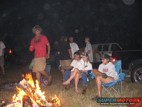 White van parked at a campsite at night with people gathered around a campfire