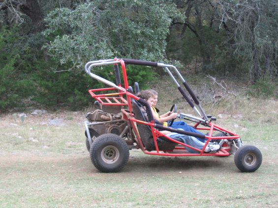 Red dune buggy with two kids driving on a grassy area with trees in the background