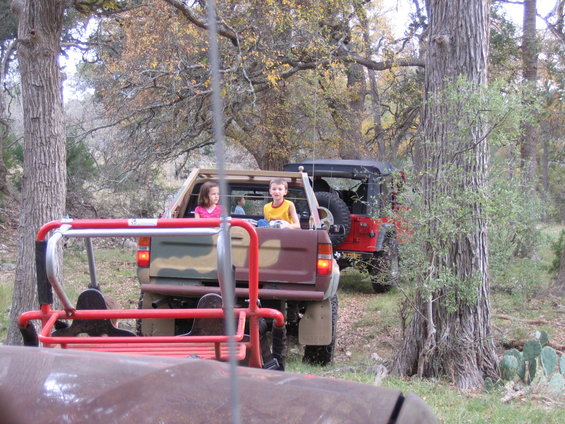 Red Jeep Wrangler and a camouflaged pickup truck with children in the back, parked in a wooded area