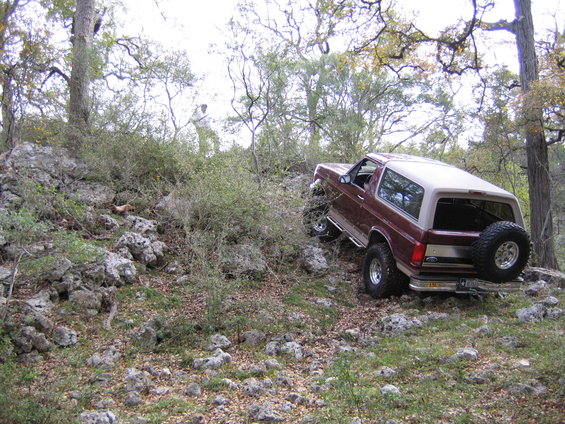 Maroon Ford Bronco with tan top navigating a steep, rocky hill off-road. Spare tire mounted on the rear