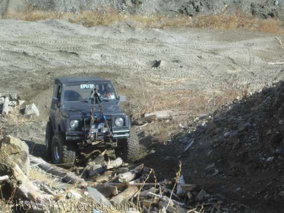 Black 1987 Suzuki Samurai driving over logs and rocks on a dirt trail