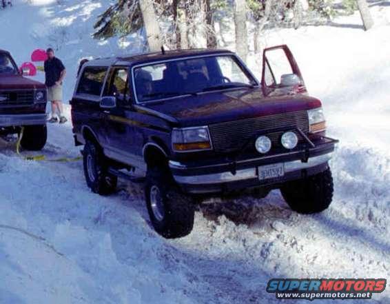 Dark blue Ford Bronco in snow with driver's door open. Red truck in background