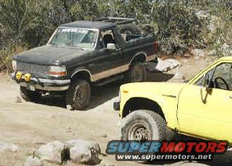 Black 1992 Ford Bronco and yellow pickup truck on a dirt road
