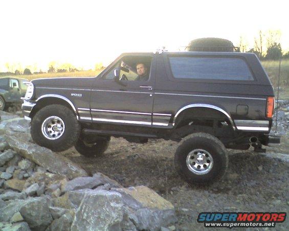 1995 Ford Bronco climbing over rocks, driver visible inside. Another SUV is in the background