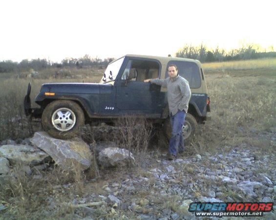 Man standing next to a dark green Jeep Wrangler with a tan top, positioned on rocks in a field
