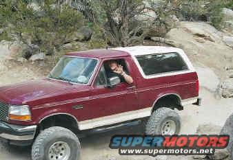 Red 1993 Ford Bronco with white top driving off-road, driver waving