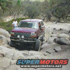 Maroon 1993 Ford Bronco navigating a rocky, off-road trail with a person standing nearby