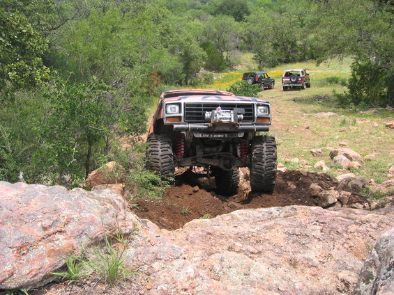 Modified 1980s truck with large tires, winch, and red suspension components navigating rocky terrain