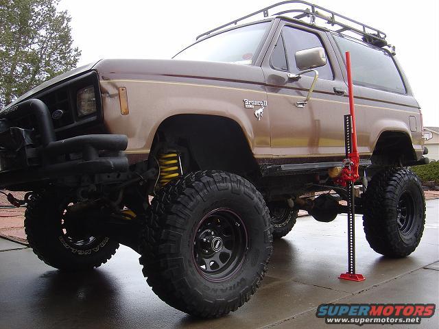 1986 Ford Bronco II, lifted with large tires, parked next to a red Hi-Lift jack