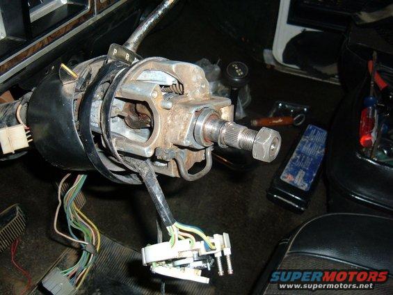Vehicle's steering column assembly with wiring harnesses disconnected. Tools and a blue rag are on the floor