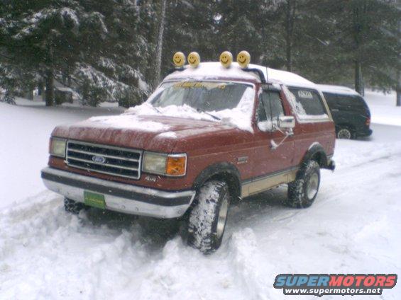 Red 1989 Ford Bronco with smiley face light covers in snow, another dark SUV behind it