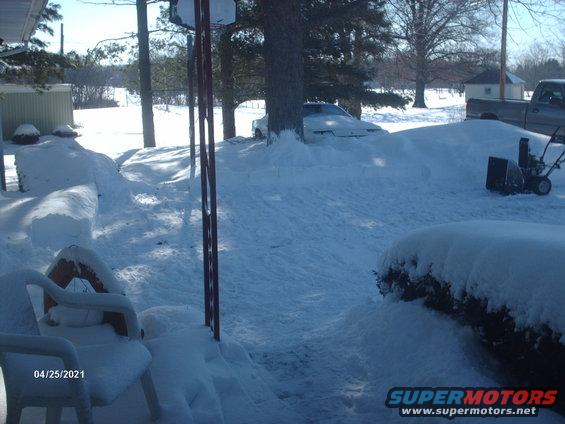 White car buried in snow, front view. A pickup truck is visible in the background