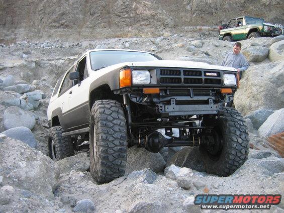 White 1985 Toyota 4Runner with large tires crawling over rocks. Another green Toyota is in the background
