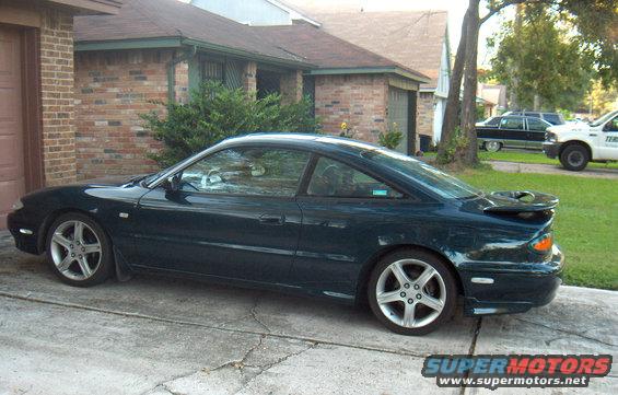 Dark teal coupe parked on a driveway, side view. It has a rear spoiler and five-spoke wheels