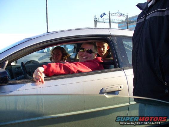 Silver car interior with three people, driver wearing sunglasses and red hoodie, passenger in back wearing glasses