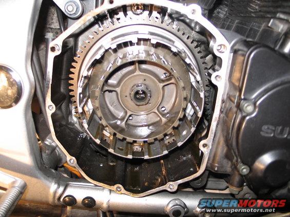Motorcycle clutch assembly with gears and plates visible. Part of engine casing is on the right with 'SUZU' visible