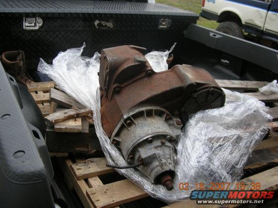 Rusty transfer case wrapped in plastic, sitting on wooden pallets in the bed of a pickup truck. Another pickup truck is visible in the background