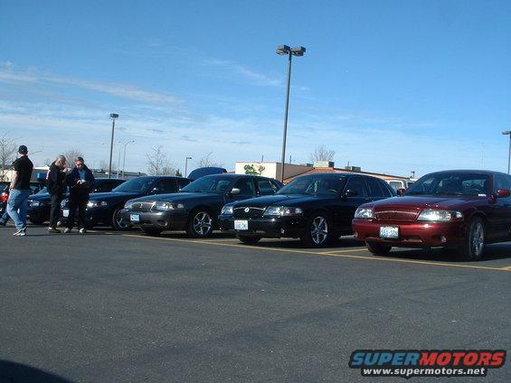 Several Ford Crown Victorias parked in a lot, one in red, others in black. Some people stand near the cars