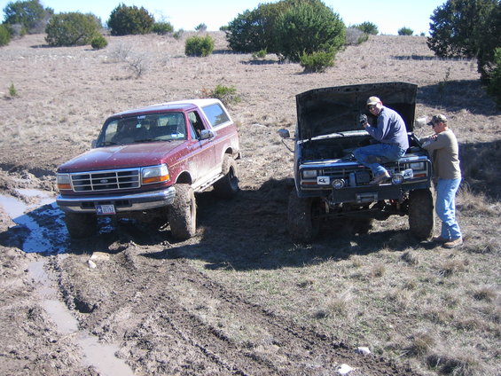 1996 Ford Bronco and another off-road truck stuck in mud with two people working on the second truck