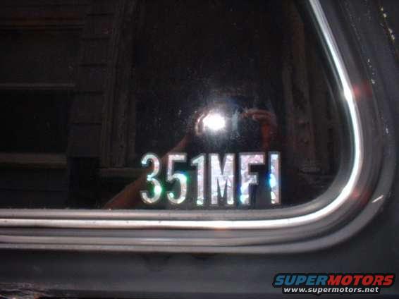Silver decal reading 351MFI on a dark window with a reflection of a camera