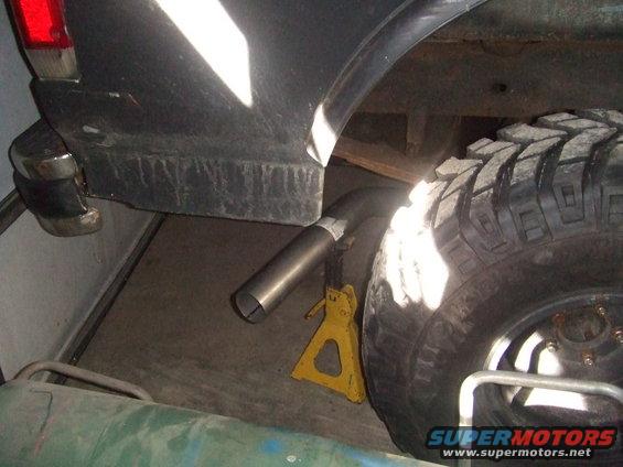 Dark colored vehicle rear quarter panel, chrome bumper, exhaust pipe, and large tire on a jack