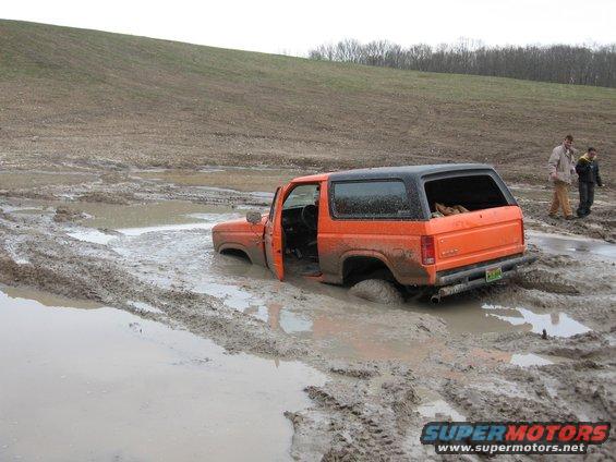 Orange Ford Bronco stuck deep in mud, driver's door open, two people standing nearby