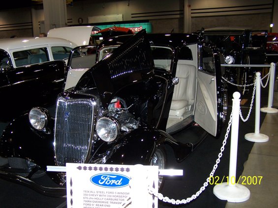 Black 1934 Ford 5-window coupe with white interior, hood open showing engine. Other cars in background