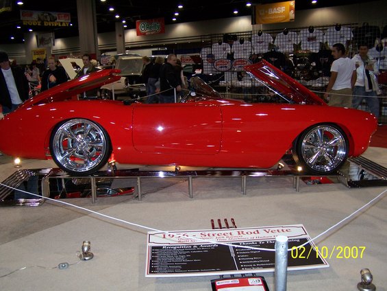 Red 1956 Corvette street rod with hood and trunk open, custom chrome wheels, on display