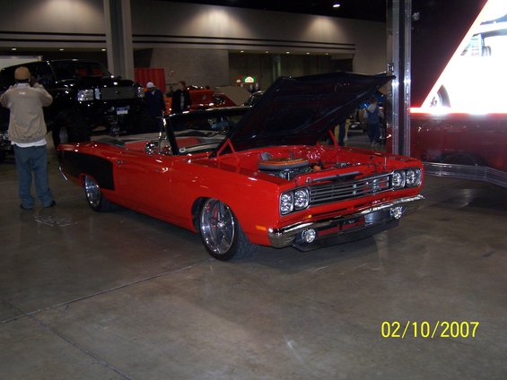 Red 1969 Plymouth Road Runner convertible with hood open, parked indoors. Black stripe on rear quarter panel. Chrome wheels