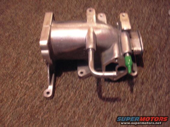 Metal intake manifold part with green plastic cap on a brown carpeted surface. Supermotors.net logo at bottom right
