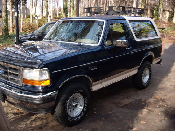 Dark blue Ford Bronco with a white top and a roof rack. Another dark vehicle is parked behind it