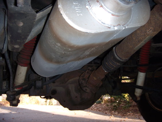 Underside view of a vehicle's exhaust, driveshaft, and rear axle. Red shocks are visible