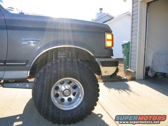 Dark gray Ford Bronco fender and tire, with a red stripe along the body. The vehicle is parked on a concrete driveway