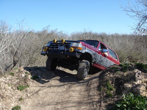 Red and grey Ford Bronco with off-road tires and yellow fog lights climbing a dirt hill