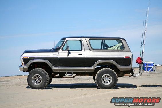 Two-tone black and silver Ford Bronco on a sandy beach, side view. Fishing rods are mounted on the rear