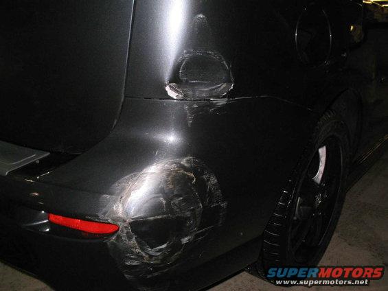 Rear of a dark-colored vehicle with significant damage to the quarter panel and bumper. Fuel door is visible