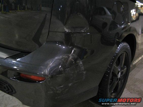 Black minivan rear bumper with significant damage, dented and scraped. Visible taillight and wheel
