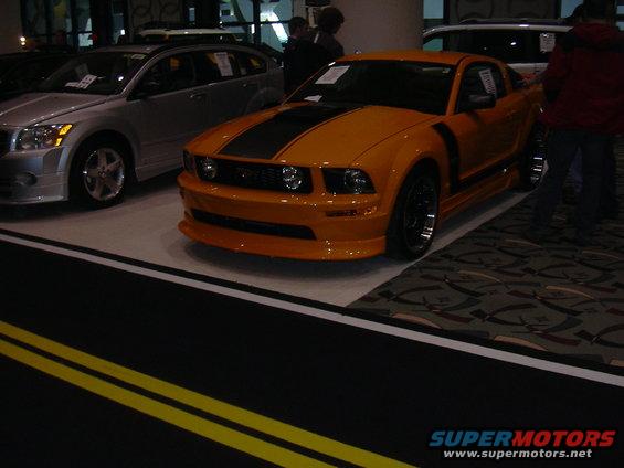 Orange Ford Mustang with black racing stripes, front 3/4 view, on display