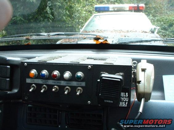 Police car interior showing a custom switch panel with radio and microphone. A light bar is visible through the windshield