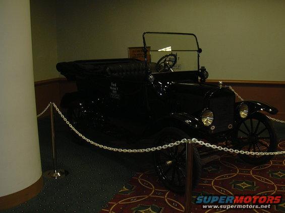 Black Ford Model T touring car with a soft top, parked indoors behind a chain barrier