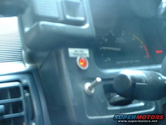 Car interior showing dashboard gauges, a red indicator light, and a toggle switch. Watermark reads SUPERMOTORS