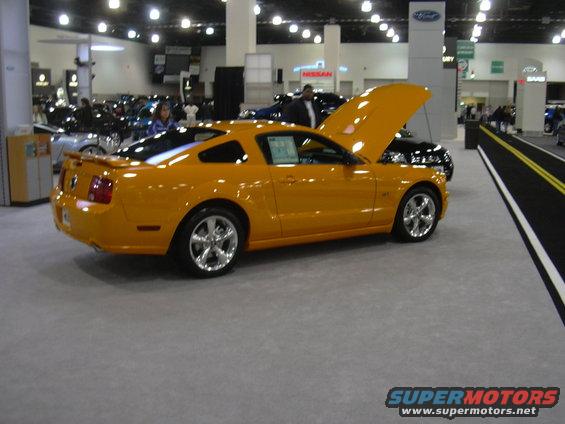 Yellow Ford Mustang coupe on display, hood open. Other cars and displays visible in background