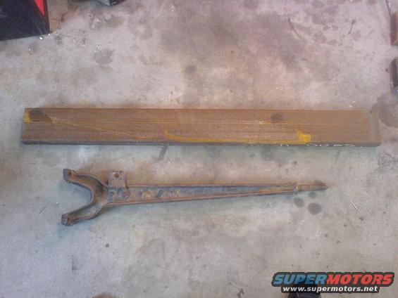 Two rusted metal components, one long rectangular bar and one curved arm with a yoke, lie on a concrete floor