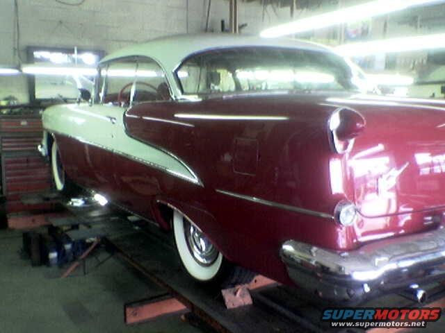 Two-tone red and white classic car on a lift, rear quarter view. Chrome bumper, whitewall tires