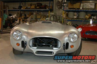 Aluminum sports car in a workshop with a red car behind it