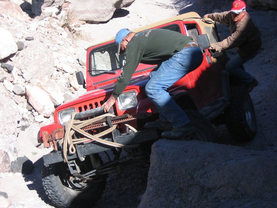 Red Jeep Wrangler stuck on rocky terrain with two people trying to free it