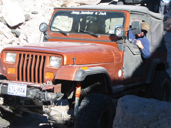Orange Jeep Wrangler with "Jeep Jambor ". JoA" sticker on windshield, driving over rocks. License plate 674-HXW
