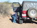 1996 Ford Bronco - A man kneels to work on the rear tire of a red Jeep Wrangler parked on a dirt road.