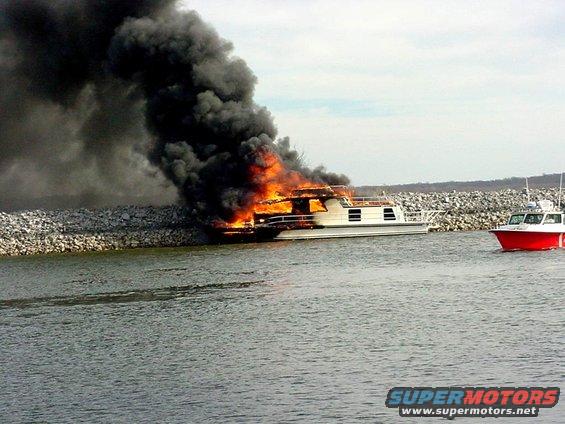 Houseboat engulfed in flames near a rock wall, with a red and white boat approaching