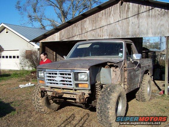 Rusty Ford pickup truck with front end damage, lifted suspension, and large tires parked under a wooden structure